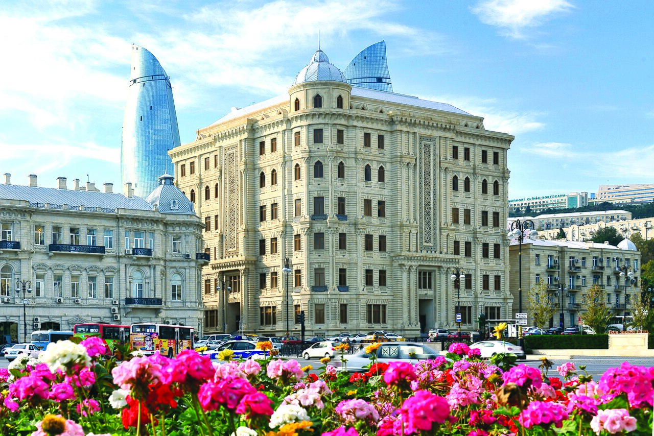 Immeubles anciens de Bakou derrière un massif fleuri, avec les tours Flame Towers en arrière-plan
