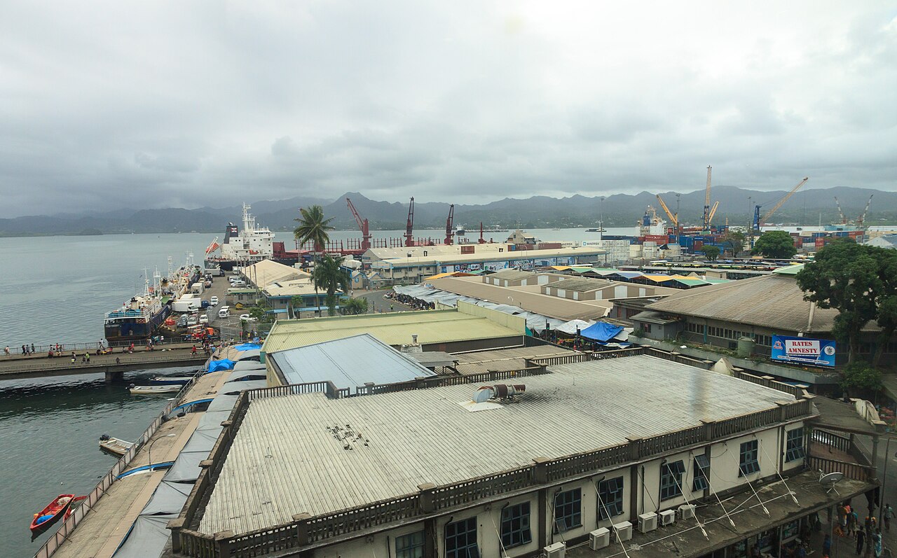 Quais et entrepôts du port de Suva avec des navires amarrés, des grues et des collines sous un ciel nuageux.